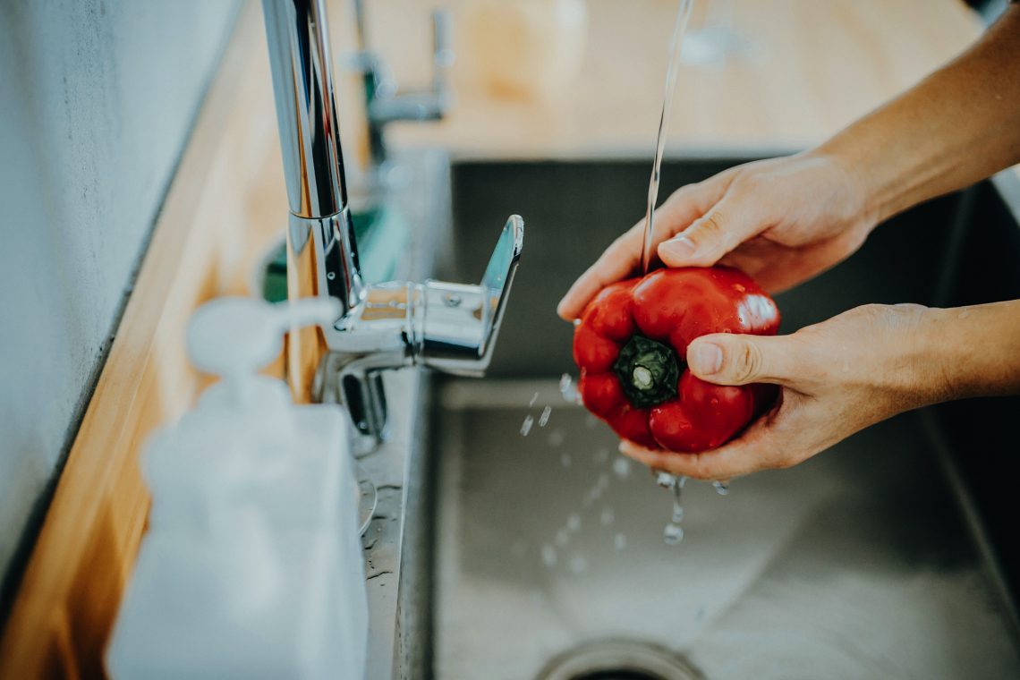 Tekst o myciu owoców, warzyw i produktów spożywczych. Na zdjęciu: Osoba myjąca czerwoną paprykę pod kranem - HelloZdrowie