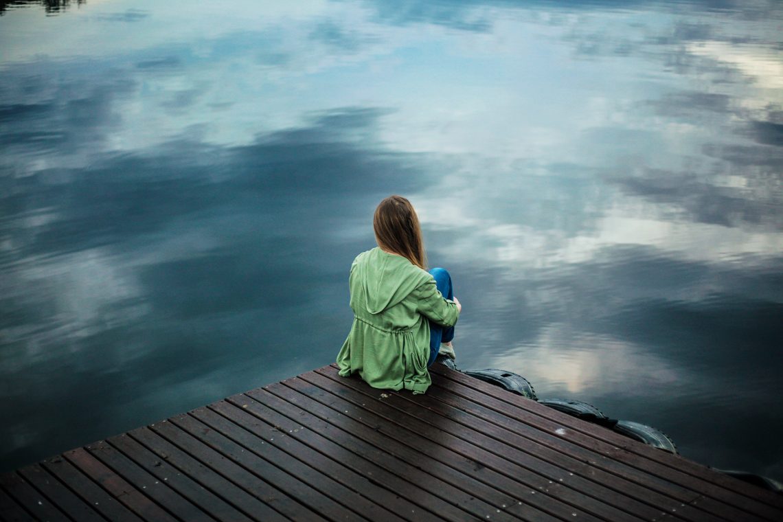 Tekst o nietypowych oznakach depresji i ich rozpoznawaniu. Na zdjęciu: Kobieta siedząca na molo i patrząca na wodę - HelloZdrowie