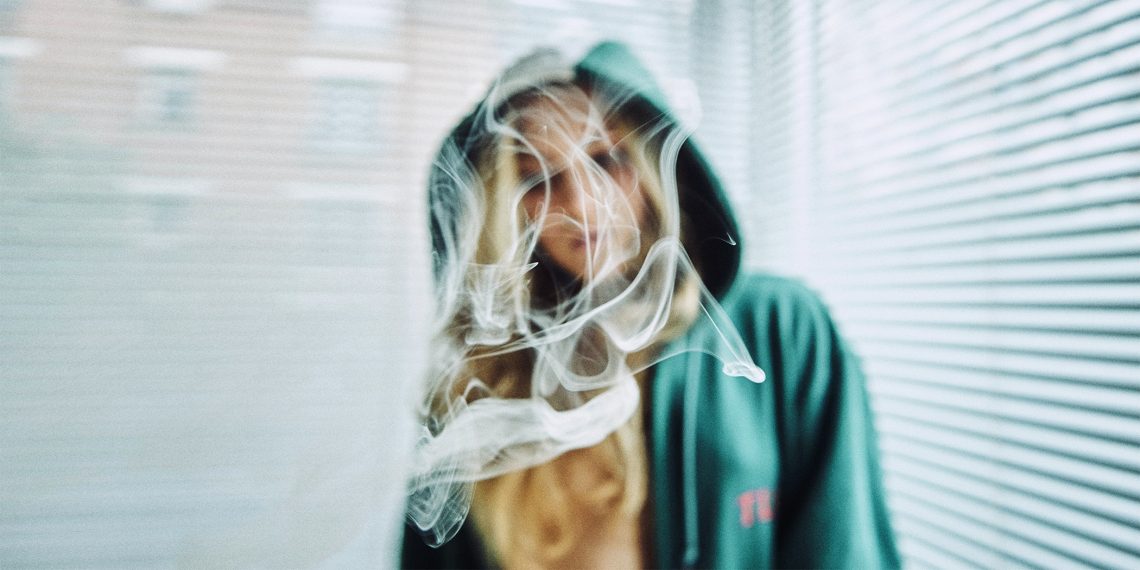 Tekst o unikaniu tycia podczas rzucania palenia. Na zdjęciu: Kobieta w bluzie z kapturem z dymem wychodzącym z jej twarzy - HelloZdrowie