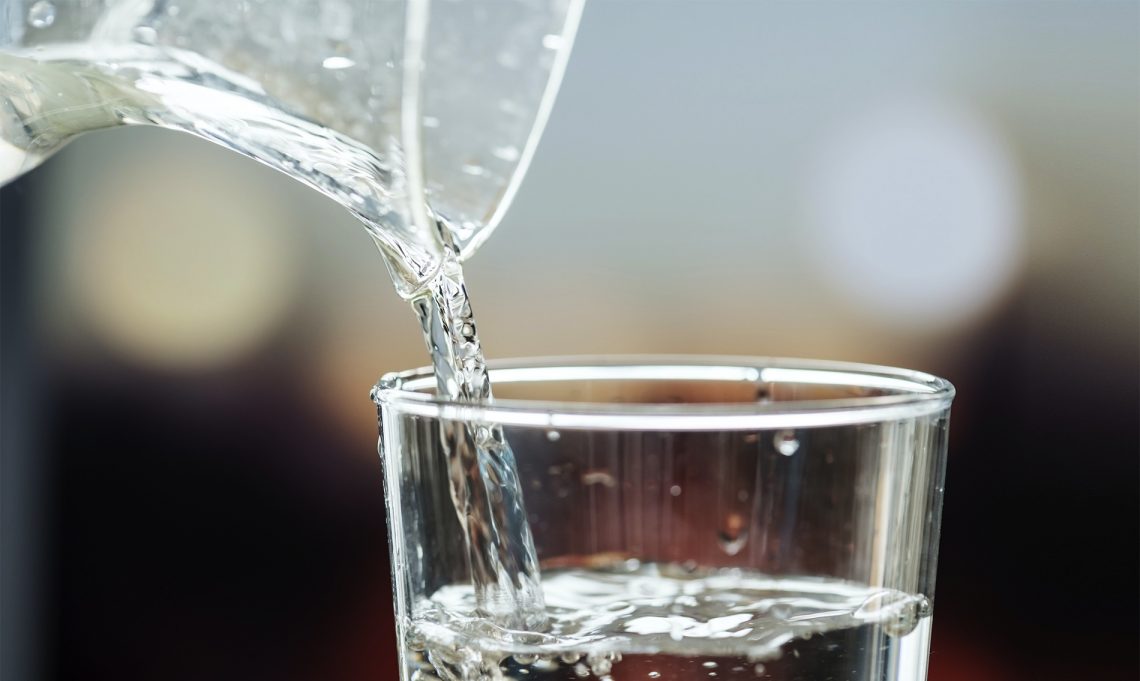 Tekst o korzyściach zdrowotnych picia ciepłej wody. Na zdjęciu: Szklanka wody wylewana do szklanki - HelloZdrowie