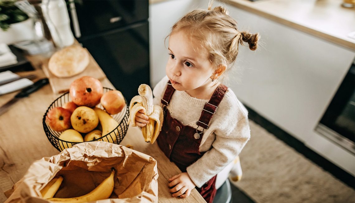 Tekst o kuchennych trikach ułatwiających życie. Na zdjęciu: Dziecko jedzące banana - HelloZdrowie