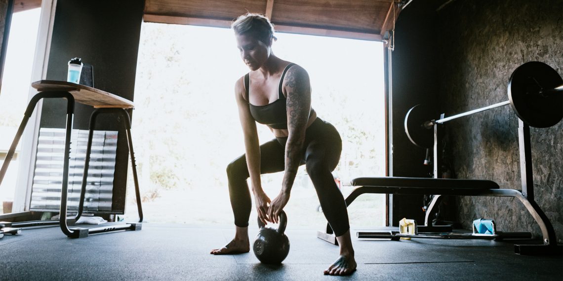 Tekst o wyposażeniu domowej siłowni podstawowym sprzętem. Na zdjęciu: Kobieta podnosząca kettlebell - HelloZdrowie