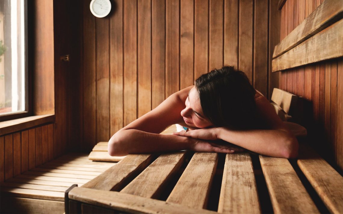 Tekst o korzyściach i zasadach korzystania z sauny. Na zdjęciu: Kobieta opierająca się o drewniany stół - HelloZdrowie