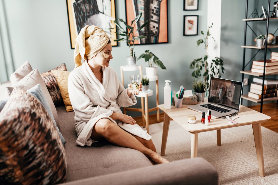 Tekst o różnicach między kosmetykami nawadniającymi i nawilżającymi. Na zdjęciu: Kobieta siedząca na sofie trzymająca szklankę płynu - HelloZdrowie