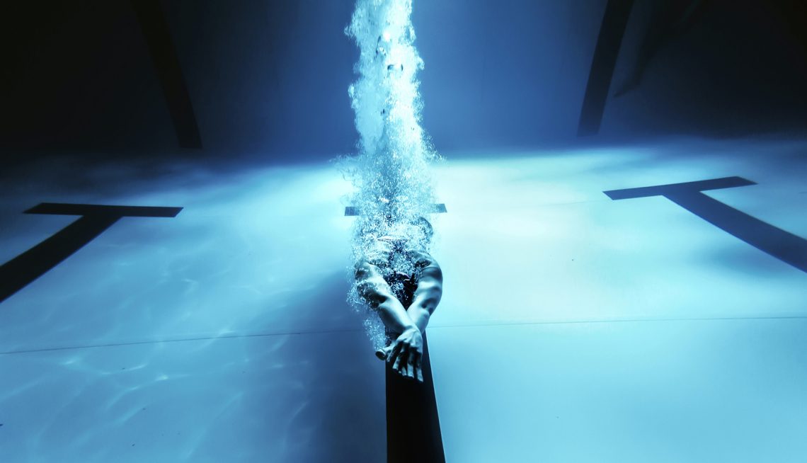 Tekst o korzyściach płynących z treningu wodnego. Na zdjęciu: Osoba pływająca pod wodą z rozpryskującą się wodą - HelloZdrowie