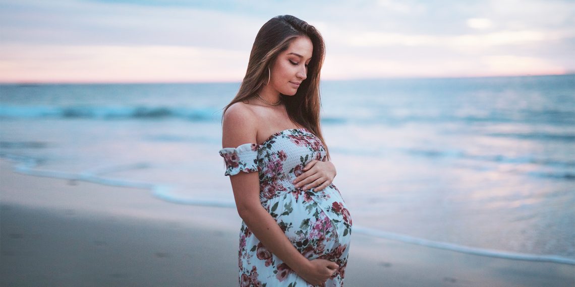 Tekst o programie Ciąża+ i jego ograniczeniach. Na zdjęciu: Kobieta w ciąży stojąca na plaży - HelloZdrowie