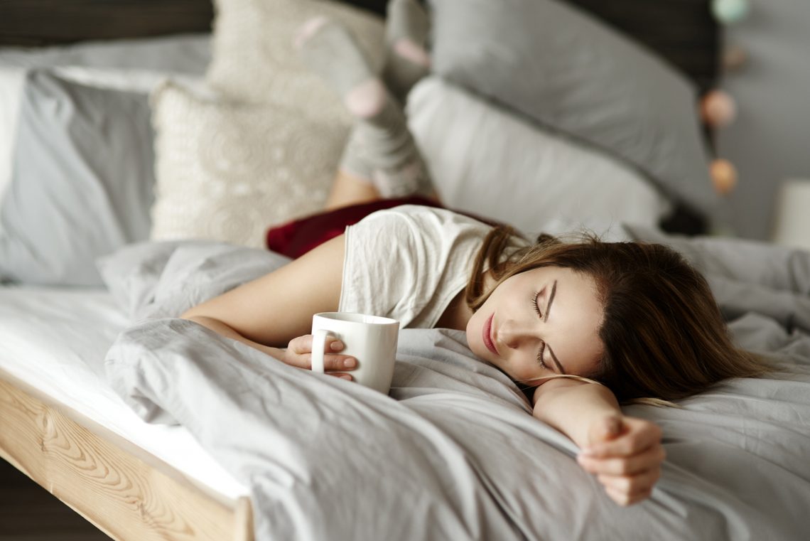 Tekst o konieczności nawadniania organizmu zimą. Na zdjęciu: Kobieta leżąca na łóżku trzymająca filiżankę - HelloZdrowie