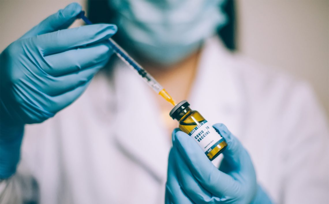 Tekst o procedurze rejestracji i szczepienia na COVID-19. Na zdjęciu: Osoba w białym płaszczu i niebieskich rękawiczkach trzymająca strzykawkę oraz butelkę z żółtym płynem - HelloZdrowie