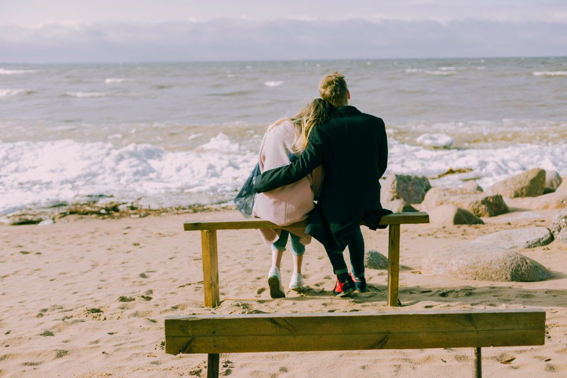 Para obejmuje się, siedząc na ławde na plaży