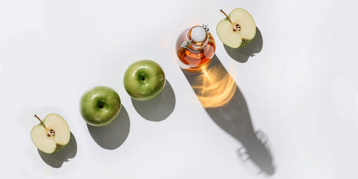 Tekst o właściwościach i stosowaniu octu jabłkowego na włosy. Na zdjęciu: Butelka soku jabłkowego obok połowy zielonych jabłek - HelloZdrowie