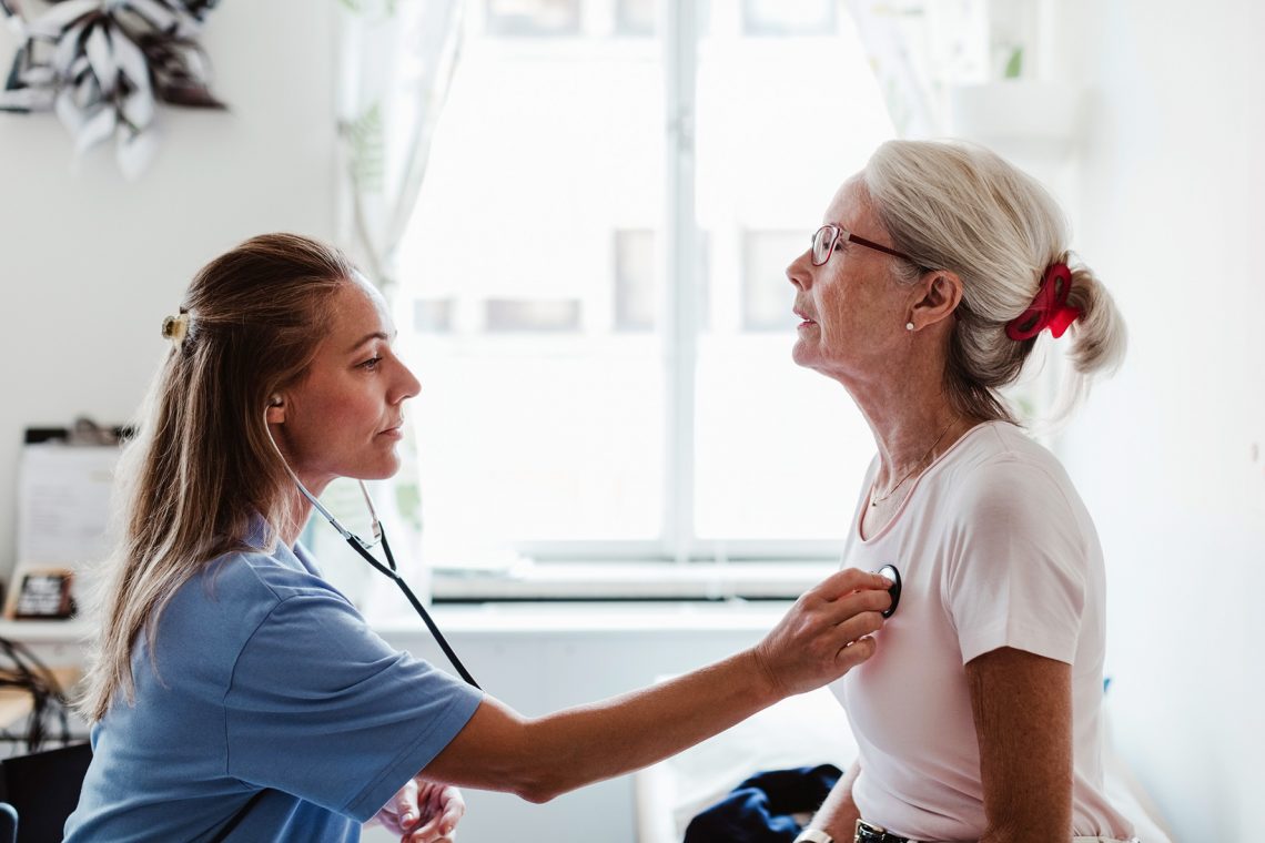 Tekst o przyczynach, typach i diagnostyce mruków serca. Na zdjęciu: Kobieta z fonendoskopem słuchająca klatkę piersiową innej kobiety - HelloZdrowie