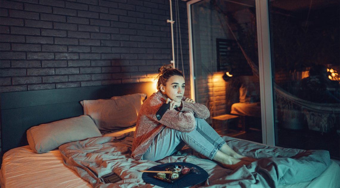 Tekst o pozytywach i negatywach binge watchingu. Na zdjęciu: Kobieta siedząca na łóżku z jedzeniem na nim - HelloZdrowie