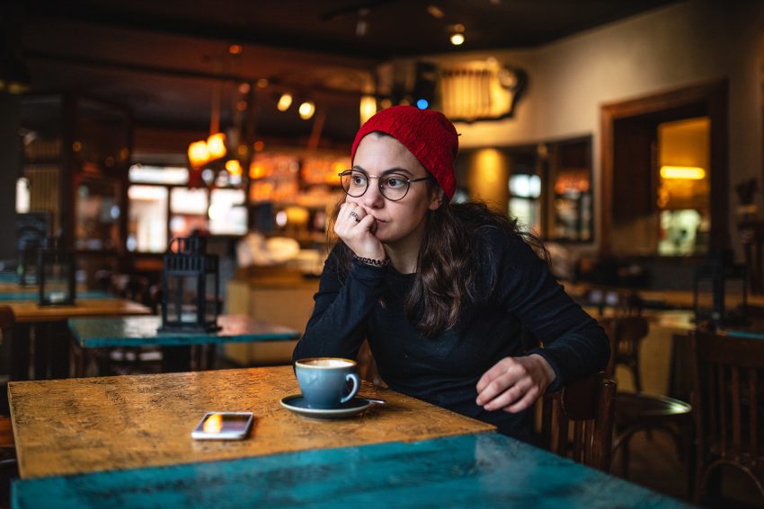Kobieta siedzi w kawiarni. Na stole soi kawa i leży telefon