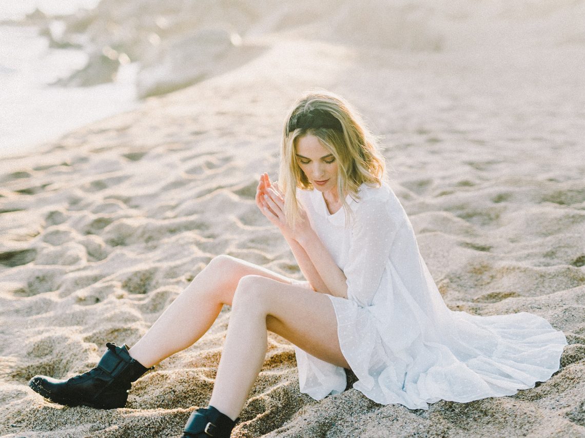 Kobieta siedzi na plaży. Ubrana jest w sukienkę i botki