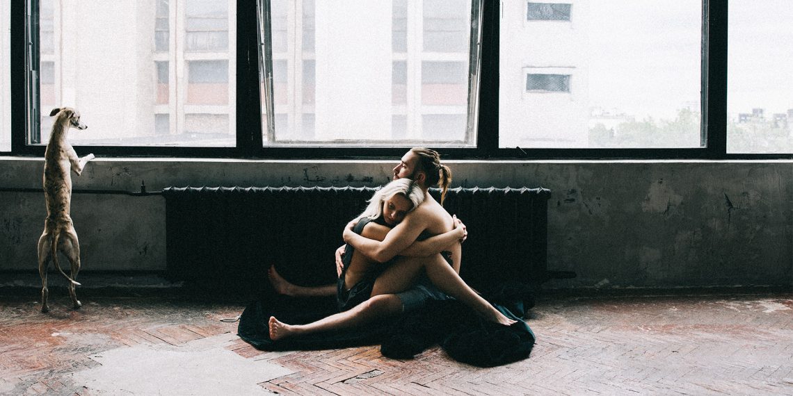 Kobieta i mężczyzna siedzą na podłodze, przytulają się. Obok stoi pies i patrzy przez okno