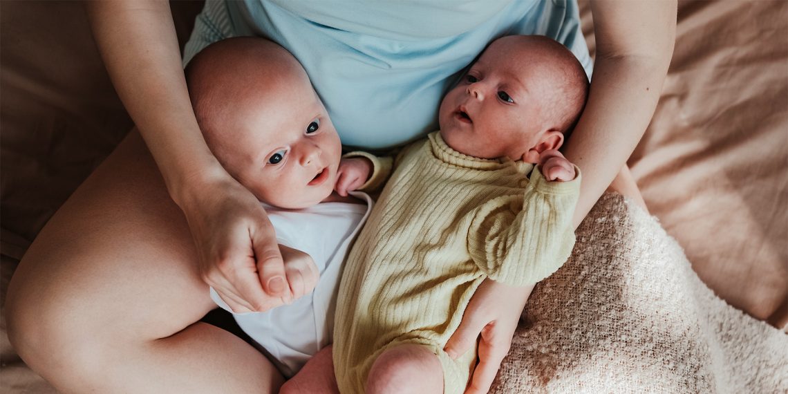 Na zdjęciu para bliźniąt- niemowląt trzymanych na kolanach przez siedzącego rodzica