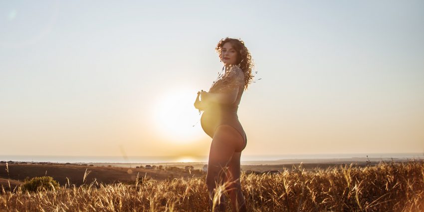 Opalanie w ciąży nie jest wskazane, ale ekspozycja na słońce już tak - na zdjęciu kobieta w ciąży w bikini stoi w trawie, w tle zachodzące słońce HelloZdrowie