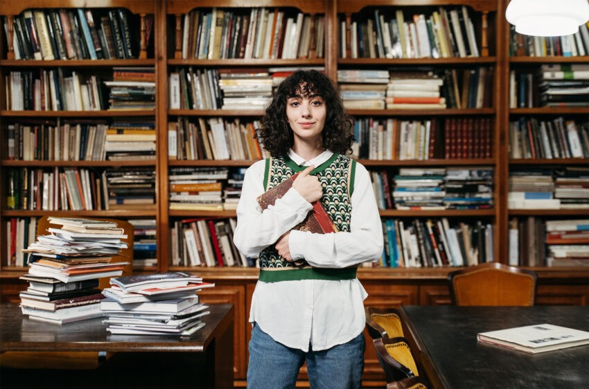 Studentka stoi w bibliotece przy regale z książkami. W rękach trzyma książkę.