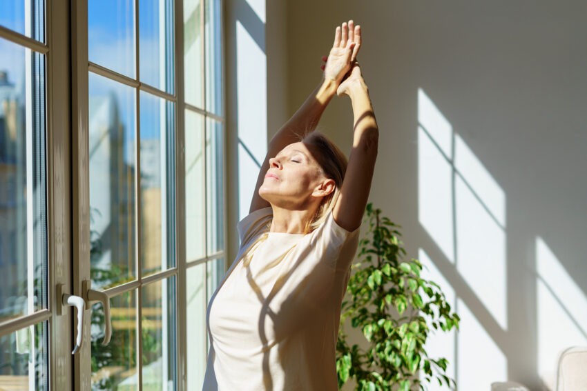 Na zdjęciu zrelaksowana kobieta przed oknem, rozciąga się- HelloZdrowie