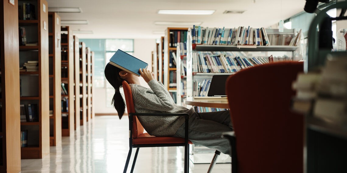 Dziewczyna siedzi w bibliotece i trzyma książkę na głowie