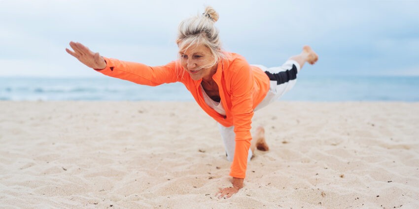 Kobieta, prawdopodobnie w okresie menopauzy, ćwiczy na plaży. Ma pomarańczową bluzę i białe spodnie dresowe, włosy związane w kok