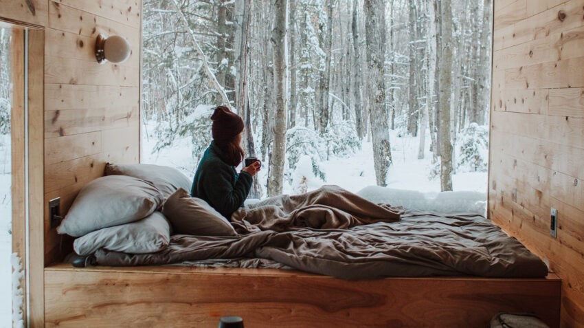 Na zdjęciu kobieta w swetrze i z kubkiem napoju obserwuje krajobraz zimowy za oknem- Hello Zdrowie