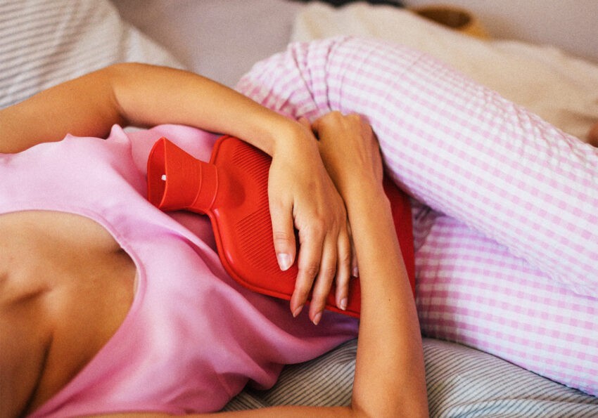 Biegunka podczas okresu. Czy powinna budzić niepokój?; na zdjęciu kobieta z termoforem na brzuchu- Hello Zdrowie