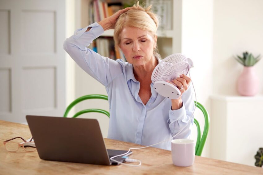 Dojrzała kobieta przy laptopie cierpi na uderzenia gorąca. Chłodzi się za pomocą wiatraka- Hello Zdrowie