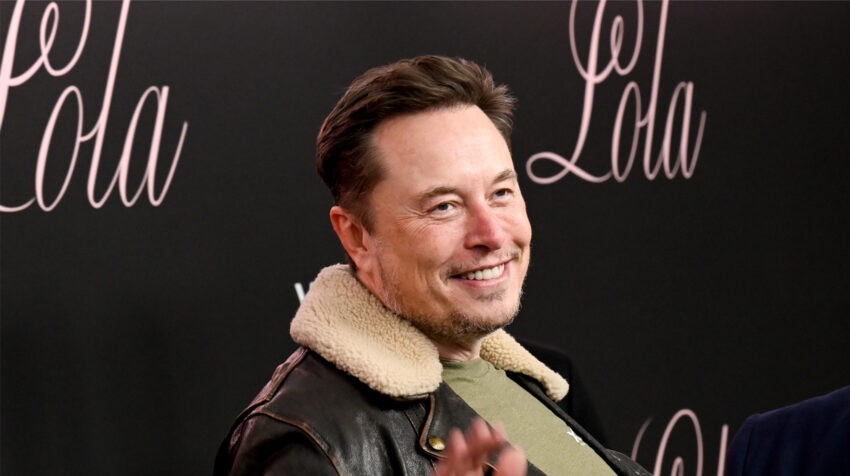 Elon Musk uśmiecha się na ściance, pozuje w skórzanej kurtce z kożuszkiem na kołnierzu