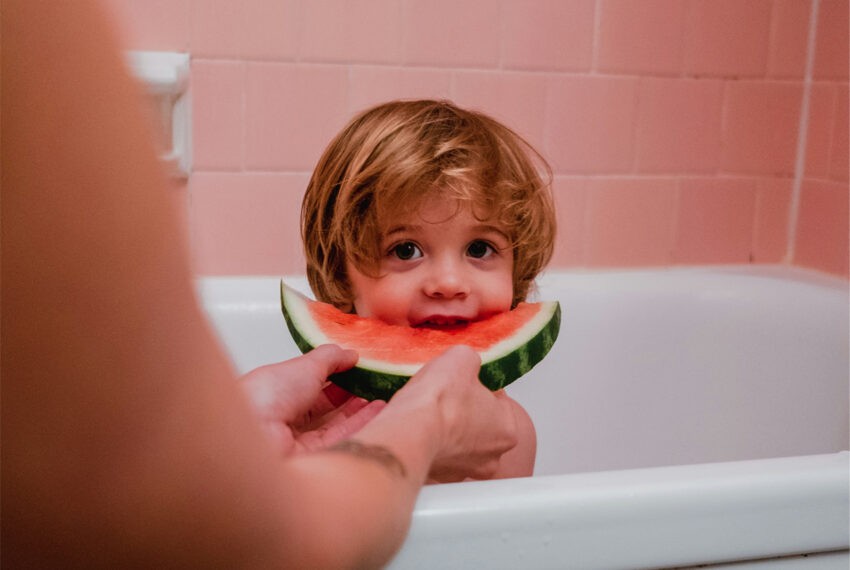 Dziecko siedzi w wannie i je arbuza podawanego mu przez mamę