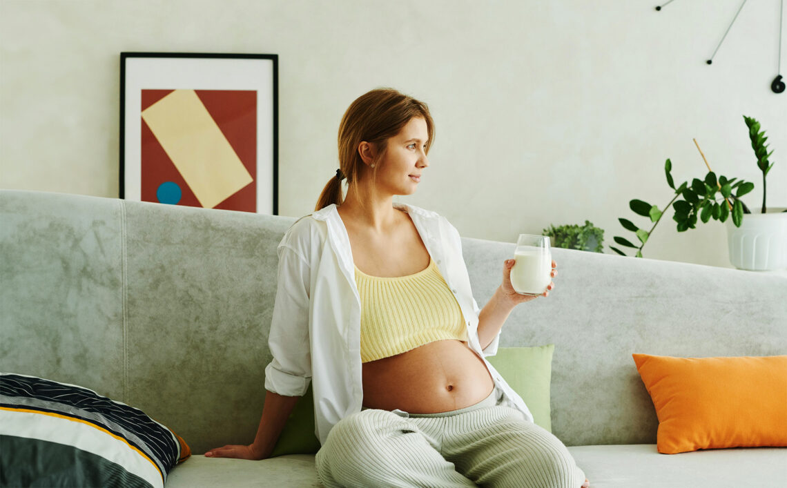 Na zdjęciu kobieta w ciąży siedząca na kanapie i trzymająca kubek z mlekiem- Hello Zdrowie