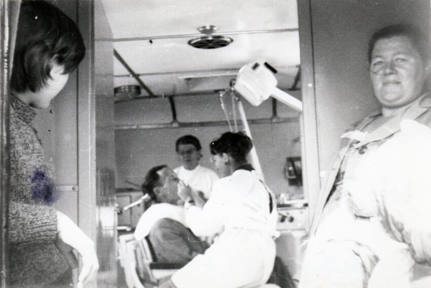 Na czarno-białym zdjęciu Teresa Radecka-Kozłowska leczy zęby pacjentowi w ambulansie dentystycznym, wszystkiemu przyglądają się dwie kobiety