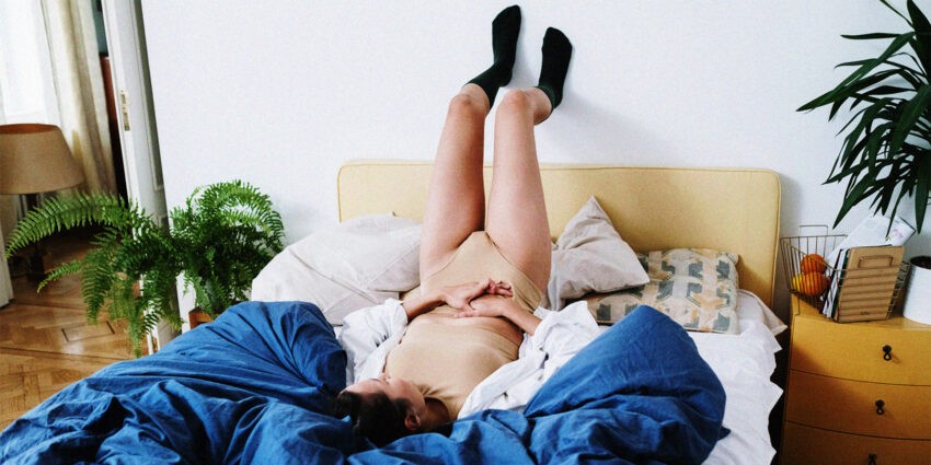 Kobieta leząca na łóżku, z nogami opartymi o ścianę. Trzyma się za brzuch\ źródło: Helllo Zdrowie