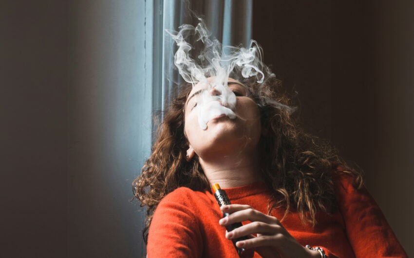 Kobieta w czerwonym sweterku i kręconych włosach wypuszcza z ust dym z e-papierosa