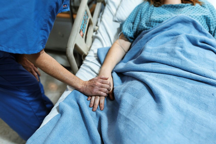 Na zdjęciu ktoś trzyma za rękę leżącą pacjentkę szpitala - Helo Zdrowie