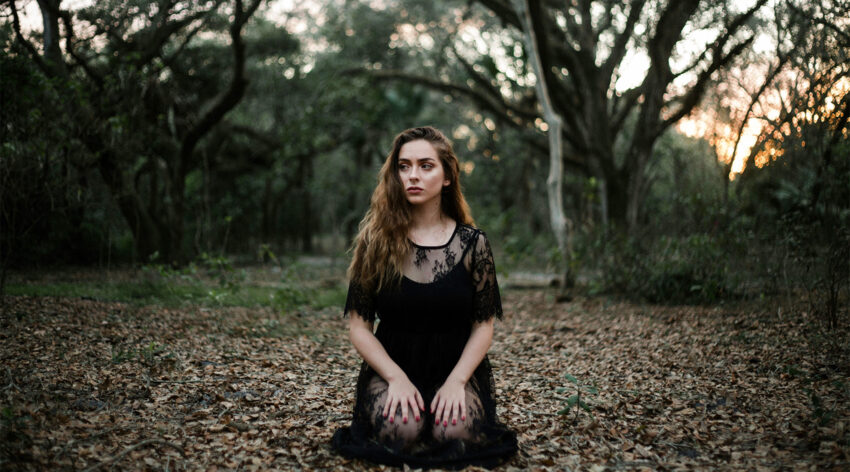 Kobieta klęczy w lesie trzymając dłonie na kolanach, ma na sobie czarną koronkową sukienkę