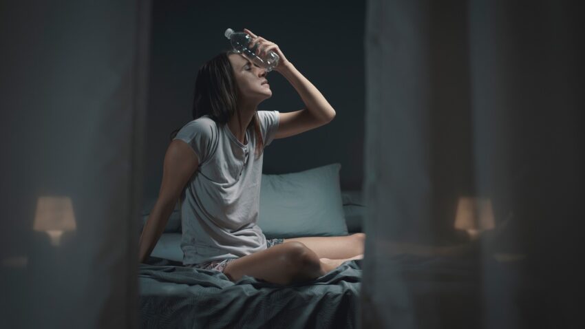 Spocona kobieta siedzi na łóżku, chłodzi się butelką wody. Tekst dotyczy nadmiernej potliwości - Hello Zdrowie