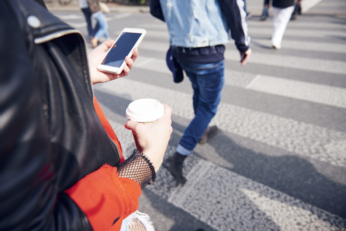 Na zdjęciu kobieta na przejściu dla pieszych z kawą i smartfonem- Hello Zdrowie