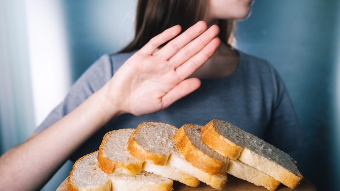 Na zdjęciu na pierwszym planie widać pokrojony chleb. Kobieta z drugiego planu wyciąga dłoń, zdecydowanie rezygnując posiłku – nietolerancja glutenu zmusiła ją do odmowy._HelloZdrowie