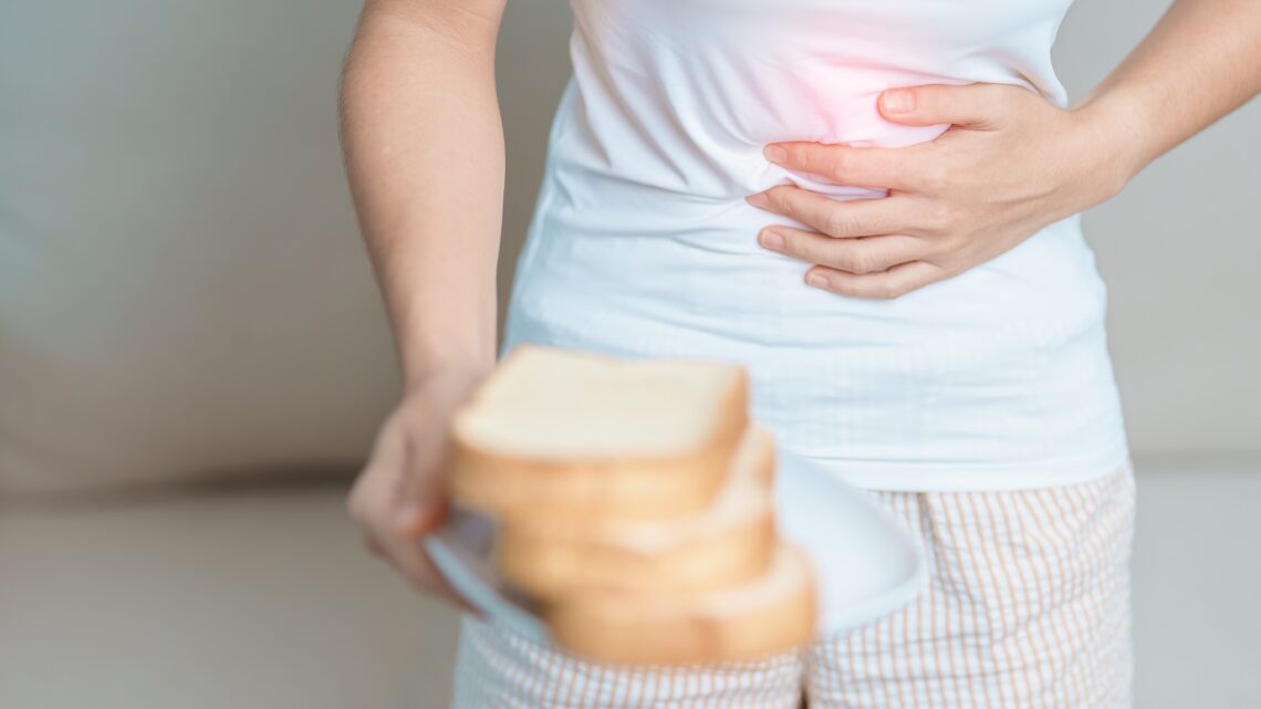 Kobieta jedną ręką trzyma kromki chleba na talerzu, drugą łapie się za bolący brzuch – to objawy nietolerancji glutenu_ Hello Zdrowie