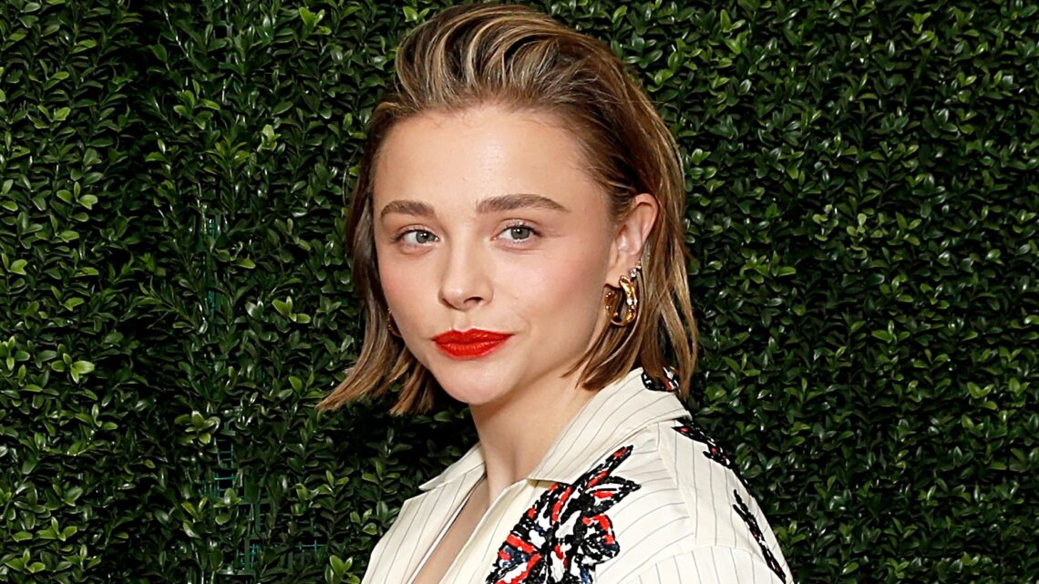 Chloë Grace Moretz pozuje na tle zielonej ścianki, ma białą marynarkę i usta pomalowane na czerwony