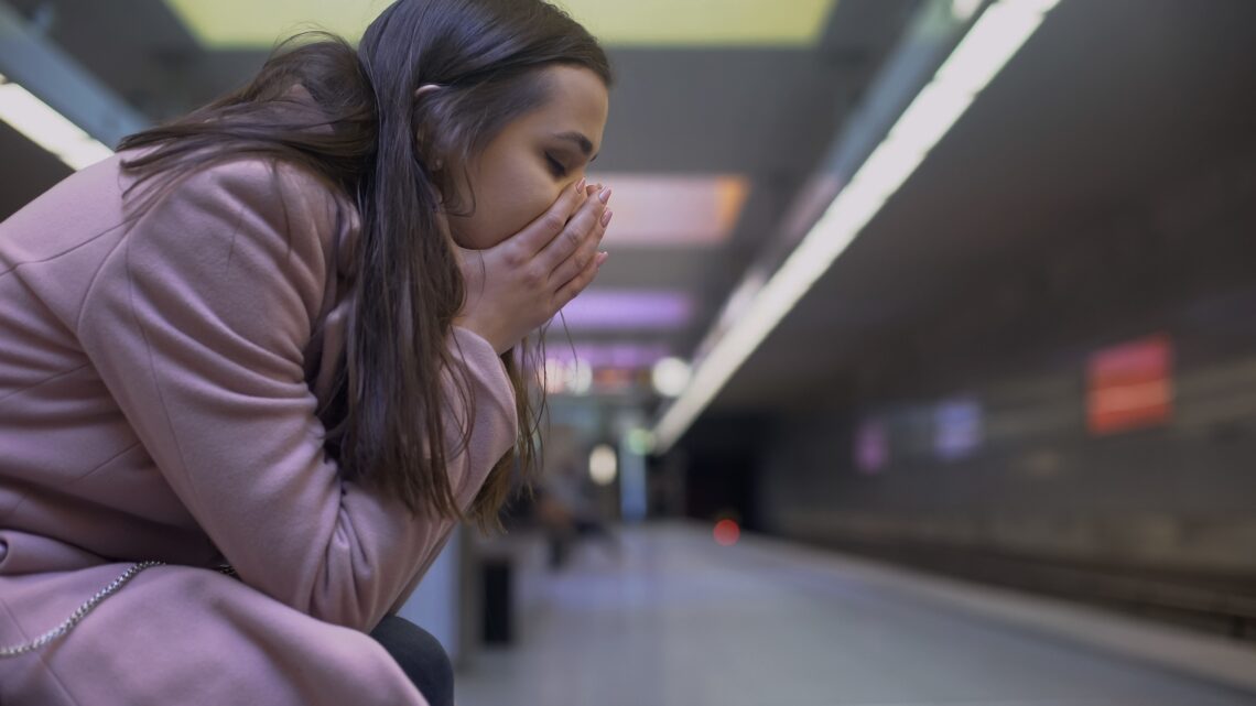 Kobieta siedzi na stacji metra, trzymając się za głowę i oddychając z trudem. Atak paniki może objawiać się nagłym uczuciem lęku, przyspieszonym oddechem, zawrotami głowy i poczuciem utraty kontroli.