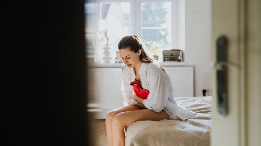 Kobieta siedzi na kanapie. Przykłada termofor do brzucha i czuje ból. Endometrioza nie jest rakiem, ale przewlekłą chorobą zapalną, która może powodować ból, zrosty i problemy z płodnością.