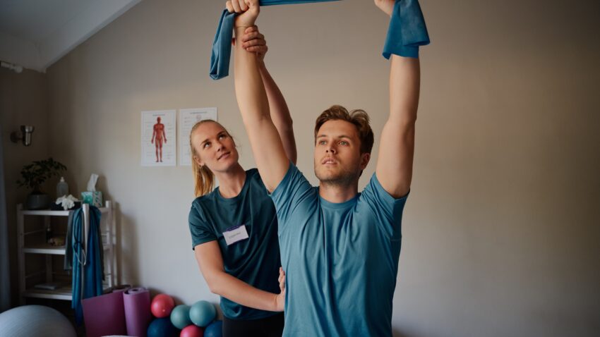 Mężczyzna wykonuje ćwiczenia z pomocą terapeuty, używając taśmy oporowej. Osteoporoza kręgosłupa może prowadzić do bólu, osłabienia kości i zwiększonego ryzyka złamań, dlatego ważna jest odpowiednia rehabilitacja i wzmacnianie mięśni.